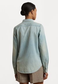 Šviesiai mėlyna džinsinė marška su smailiu apykakle, ilgomis rankovėmis ir mygtukais ant rankogalių, turinti kreivinį apačią ir subtilų malonumą.