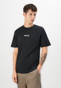 Junger Mann mit kurzen braunen Haaren, der ein schwarzes T-Shirt mit Grafik und beige Hosen trägt, zeigt ein floral und Uhr Tattoo auf seinem rechten Unterarm.