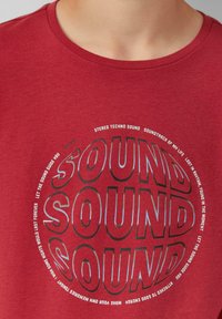 Rood katoenen t-shirt met een circulaire grafiek waarin herhaaldelijk de tekst "SOUND" in zwart en wit te zien is, omringd door verschillende muziekgerelateerde zinnen.