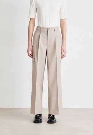 Pantalons cargo beiges avec jambes larges, devant plissé et poches latérales. Fabriqués dans un tissu lisse, associés à des chaussures noires à lacets.