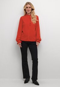 Red long-sleeve blouse with a ruffled front and button closure, paired with black flared pants and heeled ankle boots. Smooth texture.