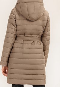 Žena v dlouhém béžovém péřovém kabátu s vodorovným prošíváním, kapucí a opaskem v pase, zobrazená zezadu.