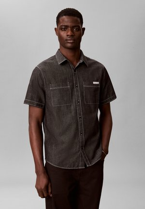 Młody mężczyzna w ciemnoszarej koszuli z krótkim rękawem i dwoma kieszeniami na piersiach, stoi na jednolitym szarym tle.