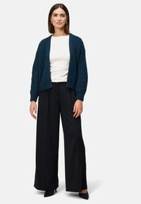 Eine Frau steht da und trägt eine dunkelblaue Strickjacke, ein weißes Oberteil, weite schwarze Hosen und schwarze High Heels vor einem schlichten hellen Hintergrund.