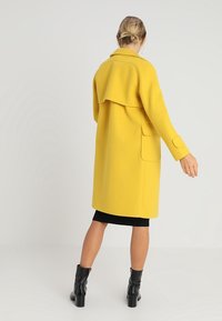 Gelber Wollmischungs-Mantel mit entspannter Passform, übergroßem Kragen und Seitentaschen. Kombiniert mit einem schwarzen Kleid und knöchellangen, glänzenden schwarzen Stiefeln.
