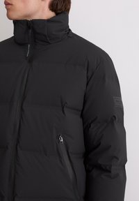 Schwarze Daunenjacke mit hohem Kragen, ausgestattet mit einem Reißverschluss vorne, Seitentaschen und einer glatten, matten Textur. Logo-Patch am Ärmel.