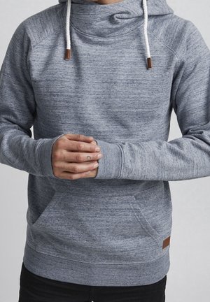 Osoba ubrana w szarą bluzę z kapturem z białymi sznurkami, z rękami złożonymi przed sobą, z małym tatuażem półksiężyca na lewej środkowej palcu.