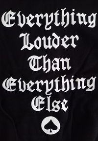 Sort t-shirt med fed hvid tekst: "Everything Louder Than Everything Else" i dekorative skrifttyper, med et kløver symbol nedenunder.