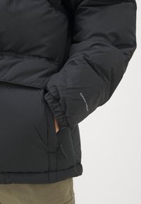 Svart boblejakke med teksturert finish, elastiske mansjetter, belte i midjen, og logo på ermet. Stoffet virker holdbart og isolert.