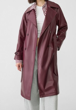 Vrouw draagt een lange bordeauxrode leren trenchcoat over een witte crop top, gestreept shirt en grijze joggingbroek tegen een effen achtergrond.
