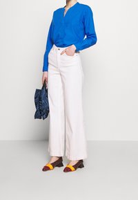 Blaues langärmliges Hemd, weiße weit geschnittene Hose und braune gemusterte Schuhe mit gelben Akzenten. Hält eine blaue Satintasche.