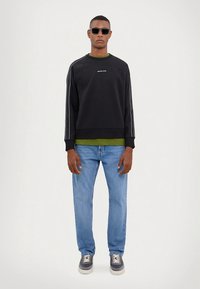 Svart genser med hvite sidestriper, grønt lag under, blå jeans og grå joggesko, båret av en person med solbriller.