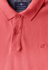 Polaire corail avec un tissu texturé, dotée d'une patte de boutonnage à trois boutons, d'un col plat et d'une broderie subtile du logo près de l'ourlet.