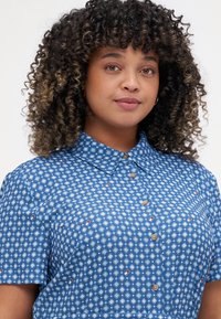 Mėlynas mygtukų marškinys su geometriniu deimanto raštu, trumpomis rankovėmis ir klasikinio tipo apykakle. Yra rudi mygtukai ir lygus paviršius.