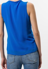 Top azul sin mangas en una tela suave y ligera con cuello fruncido. Se lleva con jeans de mezclilla claros, vista desde atrás.