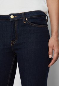 Mørkeblå denimjeans med en tettsittende design, synlig søm i gull og en metallknappstenging i midjen. Detalj av lommene foran vises.