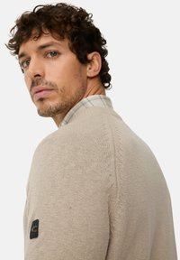 Beige knitted sweater with a textured finish, featuring a subtle design on shoulder seams and a dark logo patch on the sleeve.