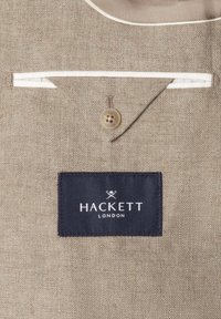 Béžová textúrovaná tkanina s bielym lemovaním, klapkou so zapínaním na gombíky a čiernou obdĺžnikovou nálepkou s nápisom "Hackett London" a logom skrížených paličiek.