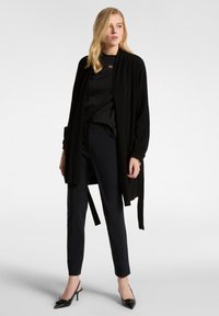 Cardigan nero, maniche lunghe, apertura frontale, abbinato a una maglietta nera e pantaloni navy sartoriali. Décolleté neri a punta completano il look.