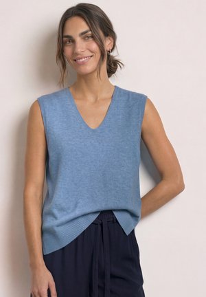 Femme aux cheveux bruns en chignon bas portant un haut en tricot sans manches bleu clair et un pantalon foncé, souriante avec une main dans le dos.