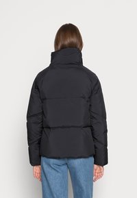 Woman with brown hair wearing a black puffer jacket and blue jeans, standing facing away against a plain white background.