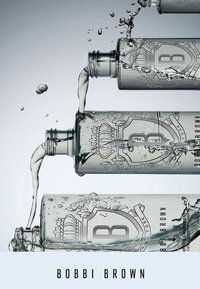 Heldere glazen flessen met reliëflogo, verschillende hoogtes, en stromende vloeistof; omringd door waterspatten op een lichte achtergrond.
