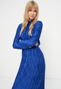 Mėlynas plisuotas suknelė su ilgomis rankovėmis ir aukštu kaklu. Audinys turi tekstūrinį, blizgantį paviršių. Tinka ties liemeniu, išsiplėtusi iki apdailos.