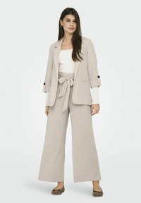 Costume beige clair avec un blazer cintré et un pantalon large, doté d'une taille ceinturée, associé à un haut blanc et des chaussures plates noires.