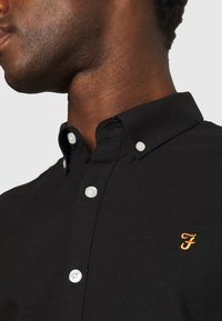 Black button-down shirt made of fabric with a smooth texture; features white buttons and a small golden embroidered logo on the chest.