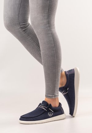 Beine in grauen, eng anliegenden Hosen, die marineblaue Slipper mit weißen Sohlen und elastischen Schnürsenkeln tragen, stehen auf weißem Hintergrund.