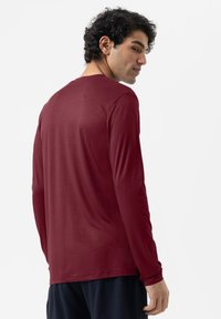 Mann mit lockigem schwarzen Haar, der ein langärmliges rotes Hemd und dunkle Hosen trägt, leicht gedreht, um die Rückseite des Hemds vor einem weißen Hintergrund zu zeigen.