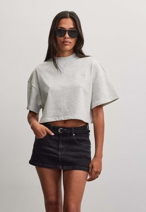 Kvinde iført sorte solbriller, lys grå kortærmet cropped sweatshirt og sort denim mininederdel med bælte, står foran en ensfarvet baggrund.