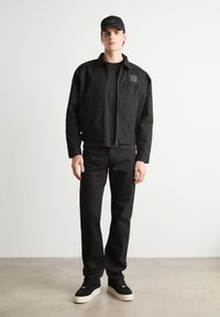 Jachetă neagră cu fermoar, țesătură texturată, tricou negru dedesubt, pantaloni negri asortați și adidași negri cu talpă albă. Capac negru purtat.
