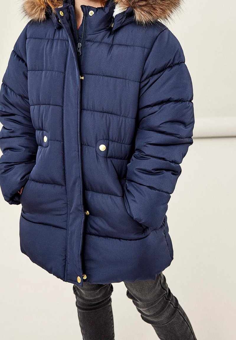 Enfant portant un manteau d'hiver matelassé bleu marine avec capuche bordée de fourrure, les mains dans les poches, debout devant un fond clair uni.