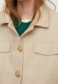 Veste en mélange de laine beige avec un col chemise, deux poches poitrine et des boutons marron, portée sur un haut côtelé vert.