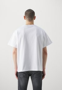 Bílé bavlněné tričko s krátkými rukávy, kulatým výstřihem a volným střihem, zobrazené ze zadní strany. Vybavené tmavě šedými džíny.