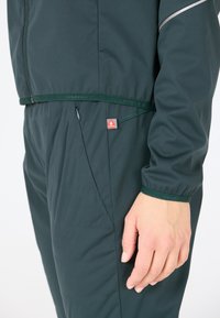 Groene waterdichte jas met rits, voorzien van een gestructureerde stof en elastische manchetten. Inclusief een klein rood logo op het taillegebied.