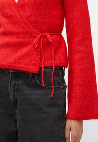 Červený pletený top s dlouhými zvonovými rukávy a nastavitelním vázáním v pase, spárovaný s tmavě šedými džínami.