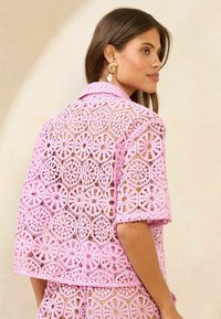 Rozā mežģīņu blūze ar sarežģītiem ģeometriskiem rakstiem, īsām piedurknēm un apkakles dizainu, izceļot puscaurspīdīgu tekstūru.