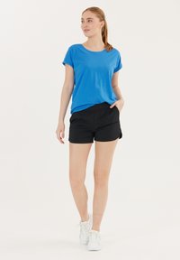 Blauwe kortemouwen T-shirt met opgerolde mouwen, gecombineerd met zwarte shorts. Model draagt witte sneakers. Eenvoudige, casual outfit met een zachte stof.