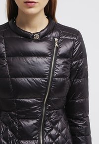 Sort dunjakke med diamantformet quiltmønster, skinnende finish, høj krave og asymmetrisk lynlåsdetaile. Detaljerne inkluderer en metaltrykknap.