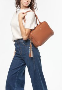 Sac à main en cuir marron de forme rectangulaire, fermeture éclair, et logo doré. Comprend un porte-clés avec un petit bijou et une étiquette.