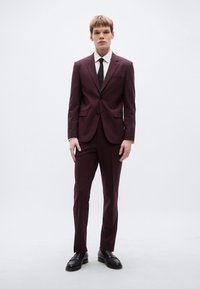 Μαρουν κοστούμι με σιλουέτα slim-fit, κλείσιμο με ένα κουμπί και πέτο με σχισμή· συνδυασμένο με λευκό πουκάμισο και μαύρη γραβάτα, φορεμένο με μαύρα παπούτσια.