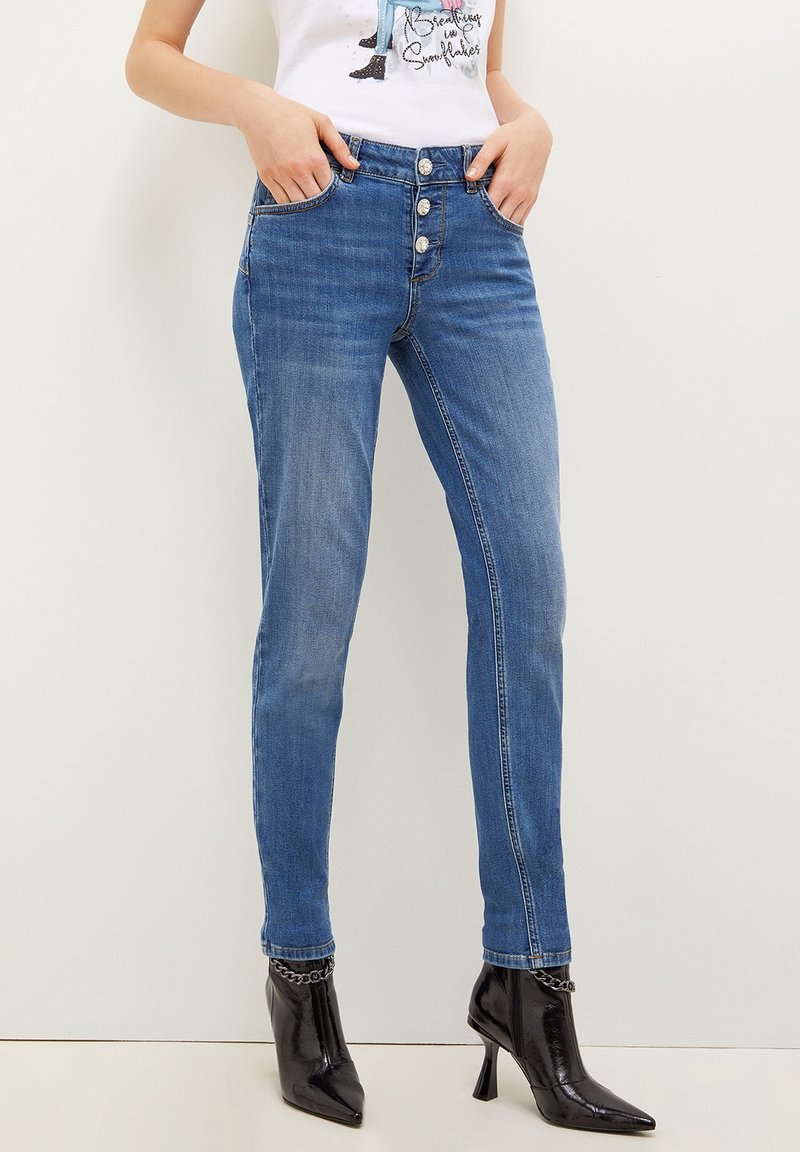 Mujer vestida con jeans ajustados de talle alto en azul medio, con tres botones frontales y botas de tacón negro de punta afilada.
