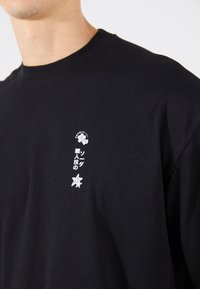 T-shirt en coton noir avec col rond. Présente des graphismes imprimés en blanc, y compris une fleur, du texte japonais et une étoile sur le côté gauche.