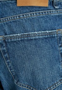Blå denimstoff med synlige sømmer, som viser en baklomme og et lærmerke på livet; teksturert finish med lett slitasje.