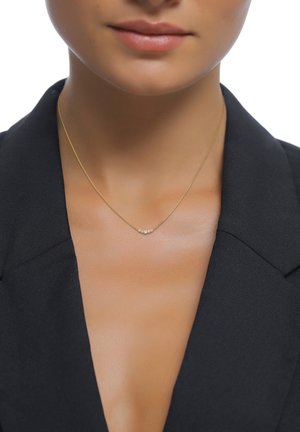 Kvinde iført en delikat gylden halskæde med små diamanter, iført en sort blazer, mod hvid baggrund.
