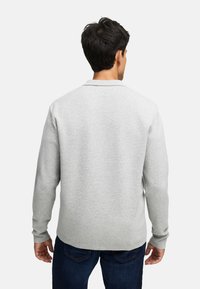 Mężczyzna odwrócony plecami, ubrany w jasnoszary, długosleeve'owy T-shirt i ciemnoniebieskie dżinsy na jednolitym białym tle.