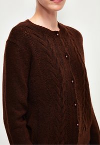 Cardigan en tricot marron présentant un motif en câble texturé, un col rond et une fermeture à boutons avec des accents ressemblant à des perles.