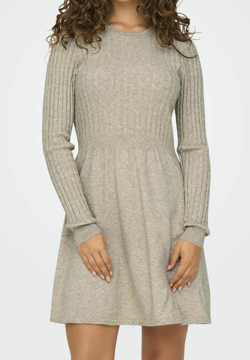 Femme portant une robe tricotée gris clair à manches longues avec des motifs côtelés et torsadés, debout les mains près de la taille.
